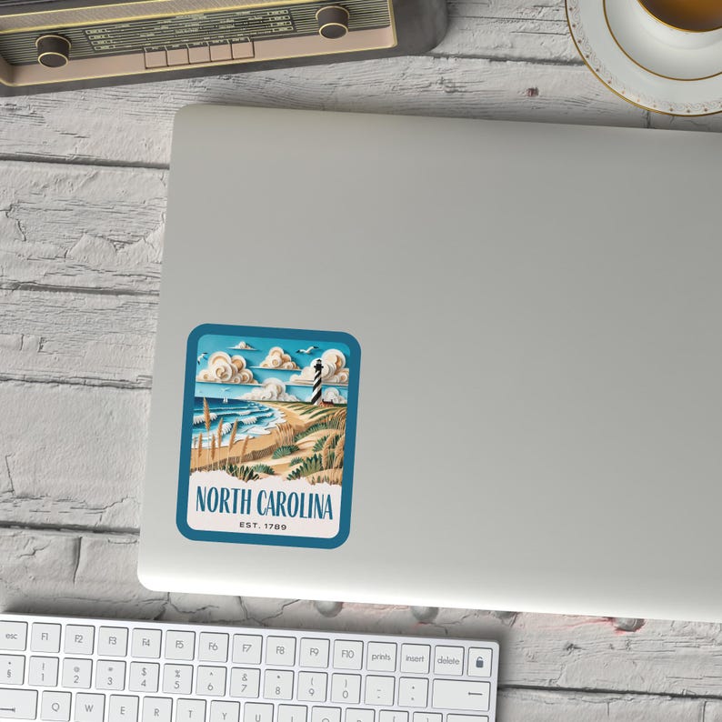 May include: A vintage-style sticker featuring a lighthouse on a beach with the text "NORTH CAROLINA EST. 1789". The sticker has a blue border and a white background.