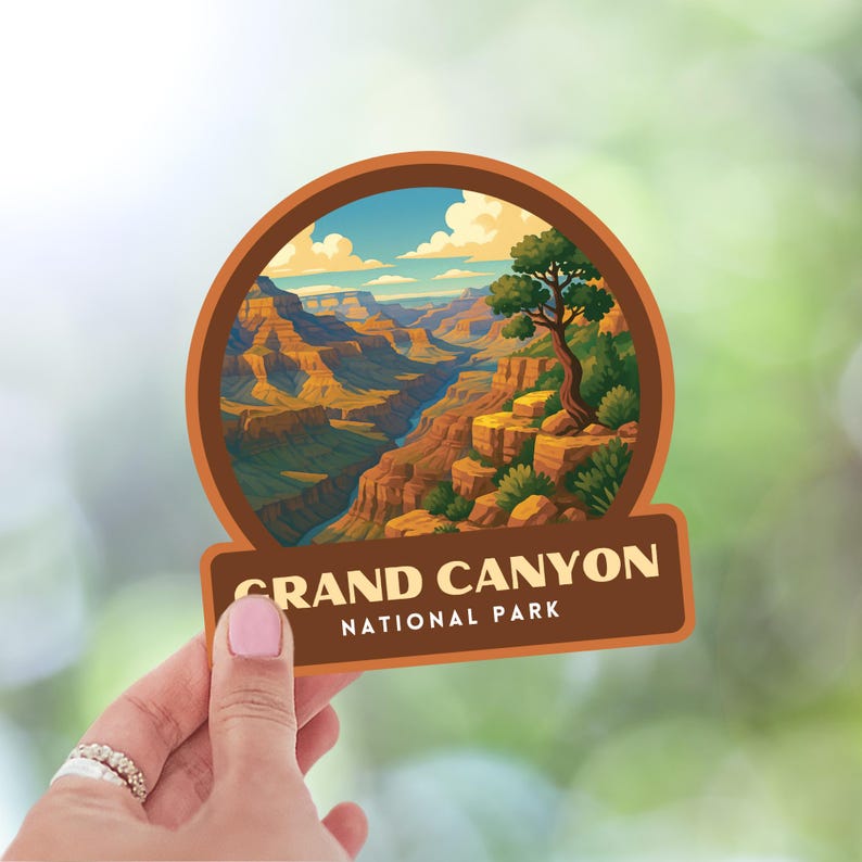 May include: A circular sticker featuring a scenic illustration of the Grand Canyon National Park. The design includes a canyon landscape with a river, a tree, and the text "GRAND CANYON NATIONAL PARK" in a brown rectangular banner.