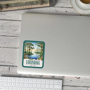 May include: A vintage-style sticker featuring a scenic view of a Louisiana bayou with cypress trees and a blue body of water. The sticker has a teal border and the text "LOUISIANA EST. 1812" in white.