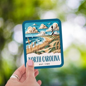 May include: A blue and white postcard with a paper cut-out illustration of a lighthouse on a beach with the text "NORTH CAROLINA EST. 1789".