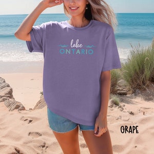 May include: A purple t-shirt with the text "lake ONTARIO" in a blue and green gradient. The text is surrounded by wavy lines. The t-shirt is being worn by a person standing on a sandy beach.