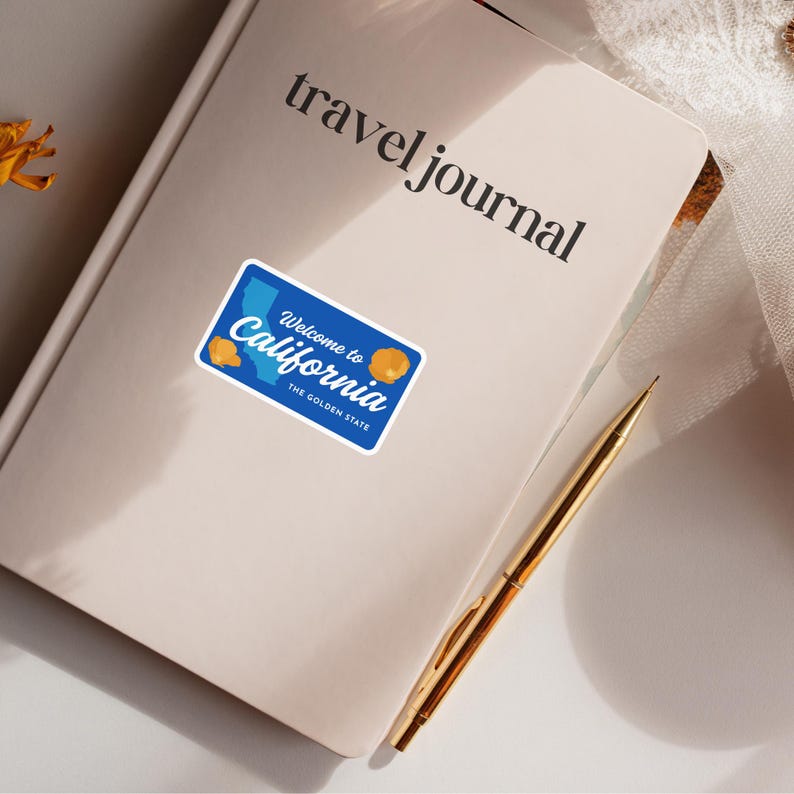 May include: A blue sticker with the text "Welcome to California" and "The Golden State" is placed on a cream-colored travel journal. The sticker features a California license plate design with yellow accents and a gold pen.