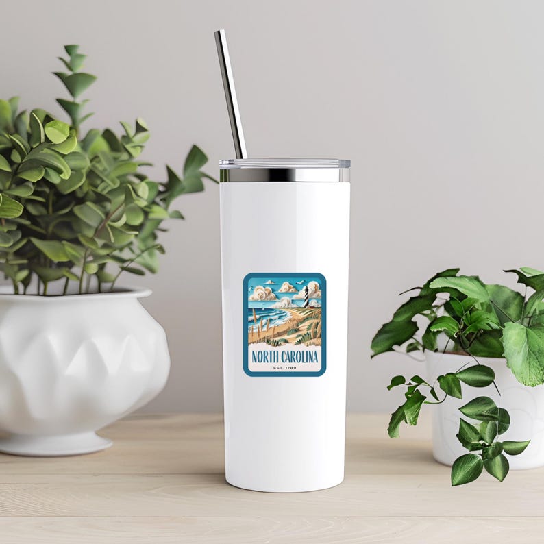 May include: White stainless steel tumbler with a blue and white sticker featuring a beach scene and the text "NORTH CAROLINA EST. 1789". The tumbler has a silver lid and a stainless steel straw.