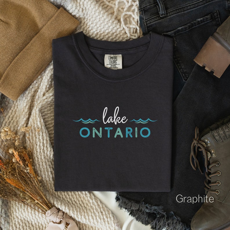 May include: A black t-shirt with the text "lake ONTARIO" in blue and green lettering. The shirt is folded and laid on a white surface with a brown hat, jeans, and brown boots.