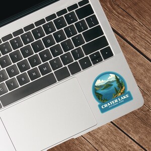 May include: A round sticker featuring a scenic view of Crater Lake National Park. The sticker shows a blue lake surrounded by green trees and mountains under a blue sky. The text "CRATER LAKE NATIONAL PARK" is displayed on the sticker.
