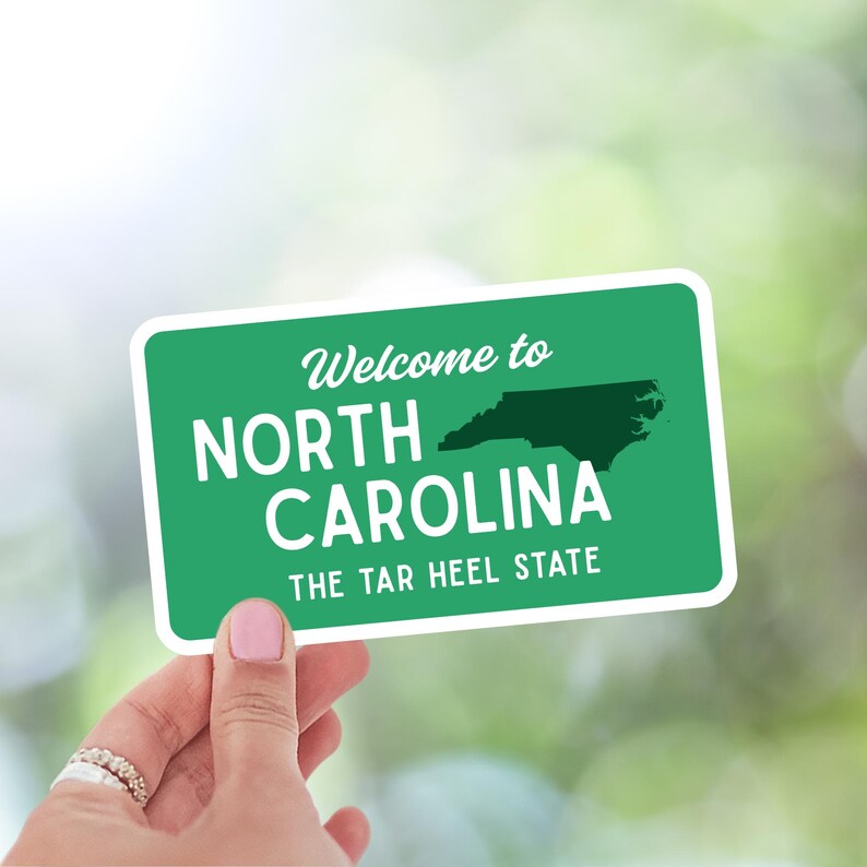 Puede incluir: Una pegatina verde y blanca con el texto "Welcome to North Carolina" y "The Tar Heel State". La pegatina presenta un contorno verde oscuro del estado. La pegatina est&aacute; sujeta por una mano.