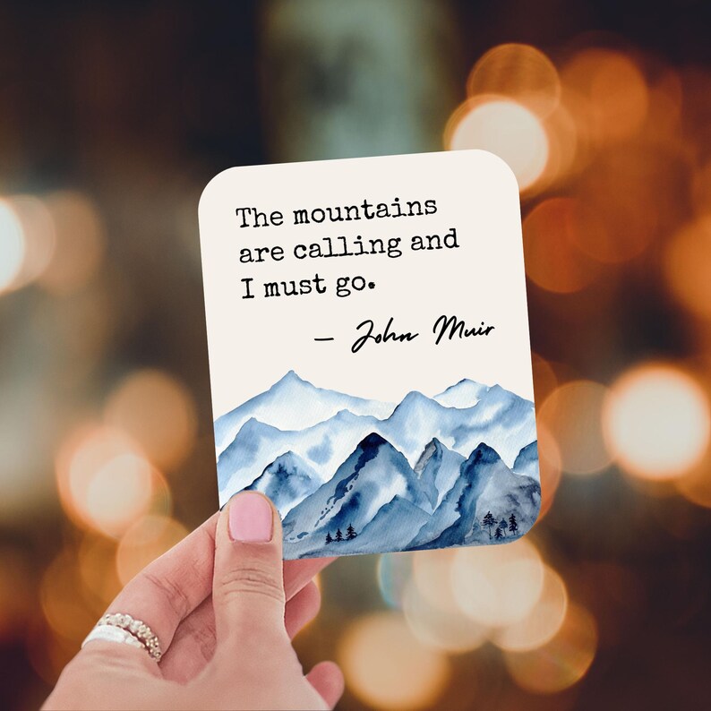 K&ouml;nnte beinhalten: Eine wei&szlig;e Karte mit einer blauen Aquarell-Berglandschaft. Die Karte zeigt das Zitat "The mountains are calling and I must go. - John Muir".
