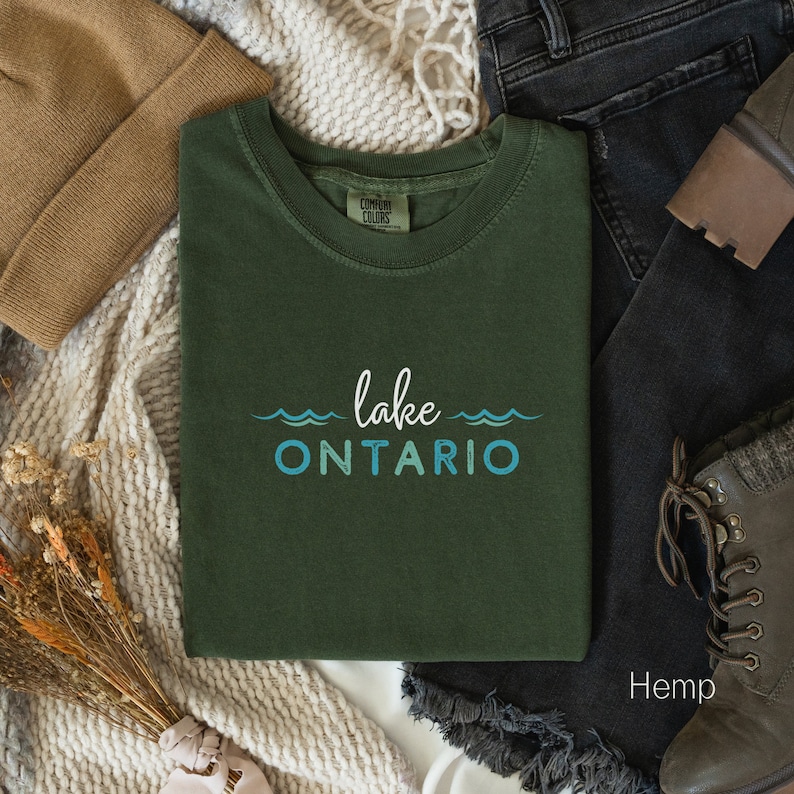May include: A green t-shirt with the text "lake ONTARIO" in blue and white lettering. The shirt is folded and laid on a white surface with a brown knitted blanket, blue jeans, and brown boots.