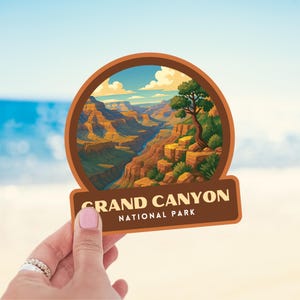 May include: A brown sticker featuring a scenic illustration of the Grand Canyon. The image shows the canyon's layered rock formations, a river, and a tree. The text "GRAND CANYON NATIONAL PARK" is displayed on a brown banner.