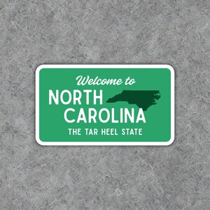 Puede incluir: Una pegatina rectangular con fondo verde y borde blanco. El texto "Welcome to NORTH CAROLINA" se muestra en blanco, con un contorno verde oscuro del estado y el texto "THE TAR HEEL STATE" debajo.