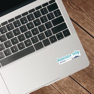 Puede incluir: Un ordenador port&aacute;til con una pegatina en el lado derecho del trackpad. La pegatina es blanca con texto azul que dice "kansas city MISSOURI | 39.0997&deg; N, 94.5786&deg; W".