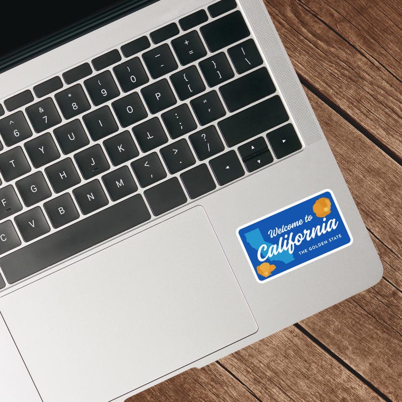 May include: A blue sticker with the text "Welcome to California" and "The Golden State." The sticker features a map of California and two orange illustrations. The sticker is placed on a laptop.