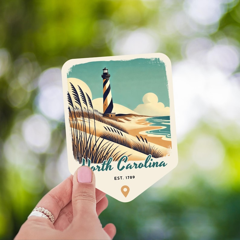 May include: A vintage-style postcard featuring a black and white lighthouse on a sandy beach with green grass and blue water. The text on the postcard reads "North Carolina EST. 1789".
