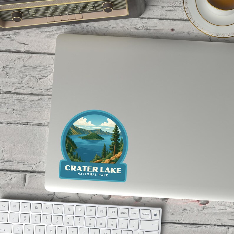 May include: A round sticker featuring a scenic view of Crater Lake National Park. The design includes a blue lake, green trees, and mountains under a blue sky. The text "CRATER LAKE NATIONAL PARK" is displayed in a rectangular banner.