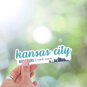 Puede incluir: Una pegatina blanca con texto azul que dice "kansas city" y "MISSOURI | N 39.10&deg;, W 94.58&deg;". Una silueta azul del horizonte de Kansas City se encuentra debajo del texto.