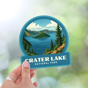 May include: A round sticker featuring a scenic illustration of Crater Lake National Park. The image shows a blue lake surrounded by green trees and mountains under a blue sky. The text "CRATER LAKE NATIONAL PARK" is displayed on a teal banner.