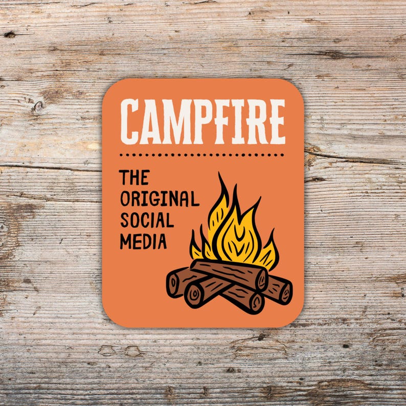 May include: Orange square coaster with the word "CAMPFIRE" in white, above the text "THE ORIGINAL SOCIAL MEDIA." A graphic of a campfire with flames and logs is below the text. The coaster is on a wooden surface.