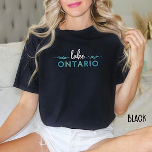 May include: A black t-shirt with the text "lake ONTARIO" in blue and green lettering. The text is surrounded by wavy lines to represent water. The t-shirt is being worn by a person with long blonde hair. The background is a white couch.