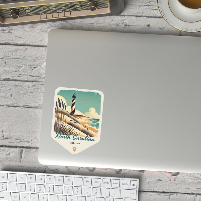 May include: A vintage-style illustration of a lighthouse on a sandy beach with tall grasses in the foreground. The text "North Carolina EST. 1789" is below the image.