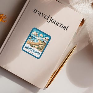May include: A travel journal with a sticker on the front. The sticker is blue and white and features a beach scene with the text "North Carolina Est. 1789".