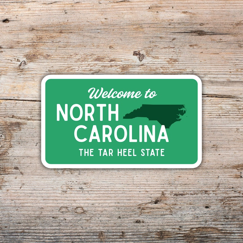 Puede incluir: Una pegatina rectangular con fondo verde y borde blanco. El texto "Welcome to NORTH CAROLINA" est&aacute; en la parte superior, con el contorno del estado y "THE TAR HEEL STATE" debajo. La pegatina est&aacute; sobre una superficie de madera.