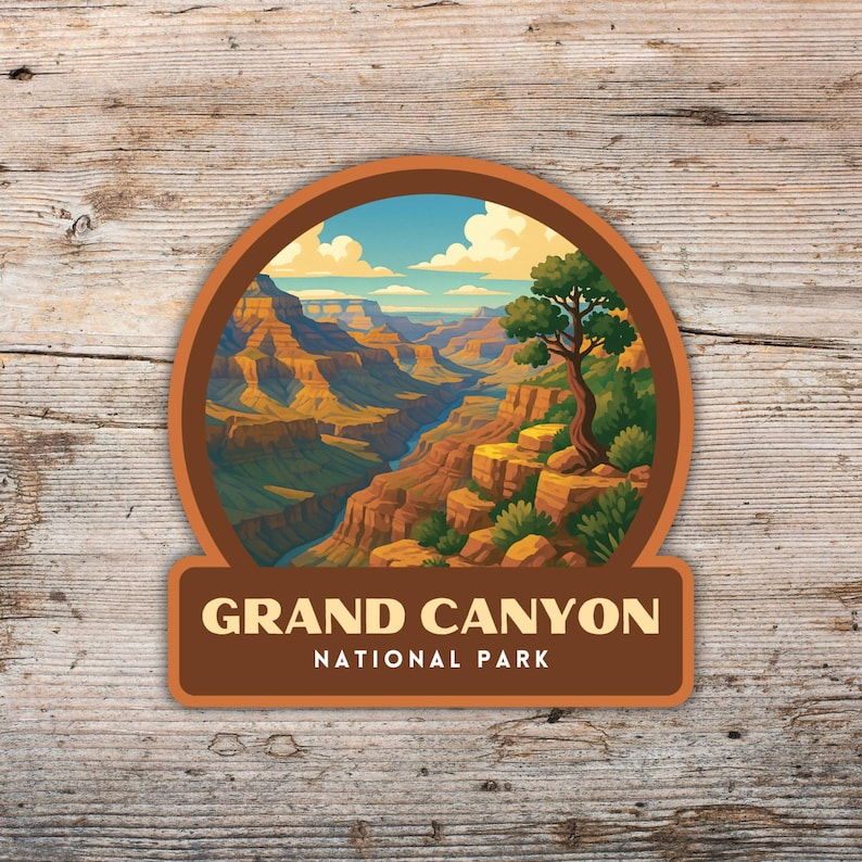 May include: A round sticker featuring a scenic illustration of the Grand Canyon National Park. The image shows the canyon's layered rock formations, a river, and a tree. The sticker has a brown border and the text "GRAND CANYON NATIONAL PARK".
