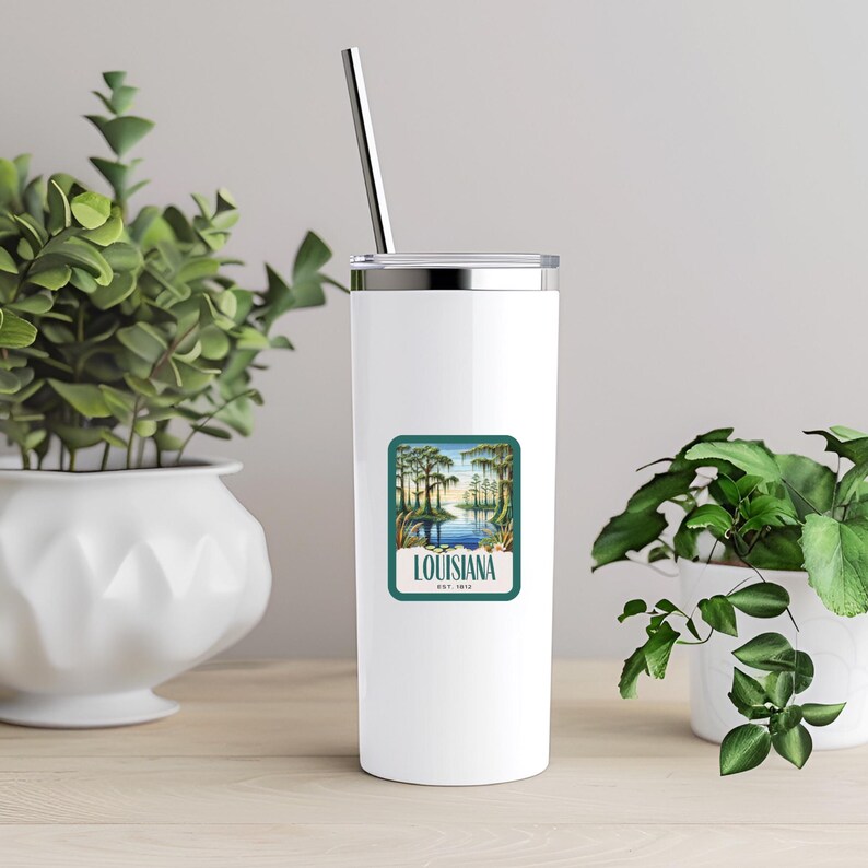 May include: White stainless steel tumbler with a green and blue sticker featuring a scenic view of a Louisiana bayou with the text "Louisiana Est. 1912".
