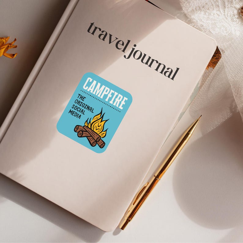 May include: A cream-colored travel journal with the words "travel journal" printed in black. A blue sticker with a campfire graphic and the words "Campfire, The Original Social Media" is affixed to the journal. A gold pen rests on the journal.