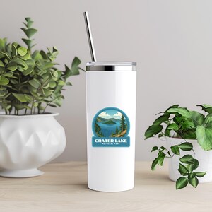May include: A white insulated tumbler with a silver lid and a metal straw. The tumbler features a circular sticker with a scenic view of Crater Lake National Park. The design includes a blue lake, green trees, and the text "Crater Lake National Park."