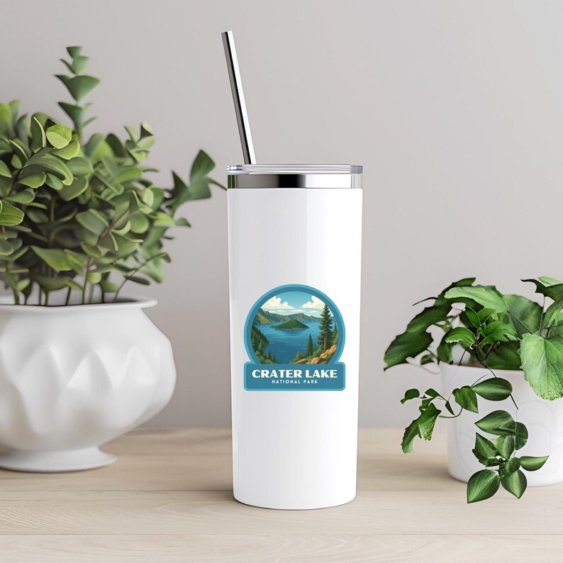 May include: A white insulated tumbler with a silver lid and a metal straw. The tumbler features a circular sticker with a scenic view of Crater Lake National Park. The design includes a blue lake, green trees, and the text "Crater Lake National Park."