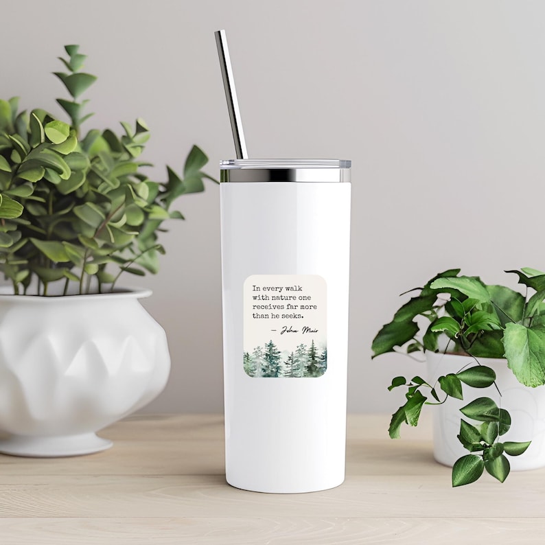 May include: White stainless steel tumbler with a quote about nature by John Muir. The quote reads "In every walk with nature one receives far more than he seeks." The tumbler has a green watercolor design of trees on the front.
