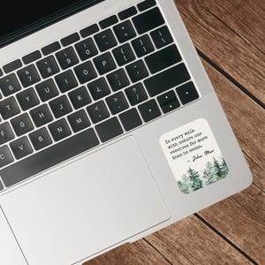 May include: A laptop computer with a sticker on the right side. The sticker features a watercolor illustration of a forest with the quote "In every walk with nature one receives far more than he seeks." - John Muir.