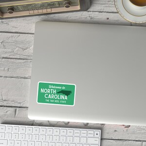 Puede incluir: Una pegatina verde y blanca con el texto "Welcome to North Carolina" y "The Tar Heel State". La pegatina es rectangular con esquinas redondeadas y presenta un contorno verde del estado.