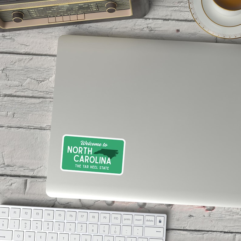 Puede incluir: Una pegatina verde y blanca con el texto "Welcome to North Carolina" y "The Tar Heel State". La pegatina es rectangular con esquinas redondeadas y presenta un contorno verde del estado.