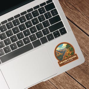 May include: A round sticker featuring a landscape of the Grand Canyon National Park. The sticker has a brown border and the text "GRAND CANYON NATIONAL PARK". The sticker is placed on a laptop.