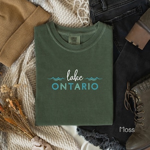 May include: A green t-shirt with the text "lake ONTARIO" in blue and white. The shirt is folded and laid on a white surface with a brown knitted blanket, brown beanie, black jeans, and brown boots.