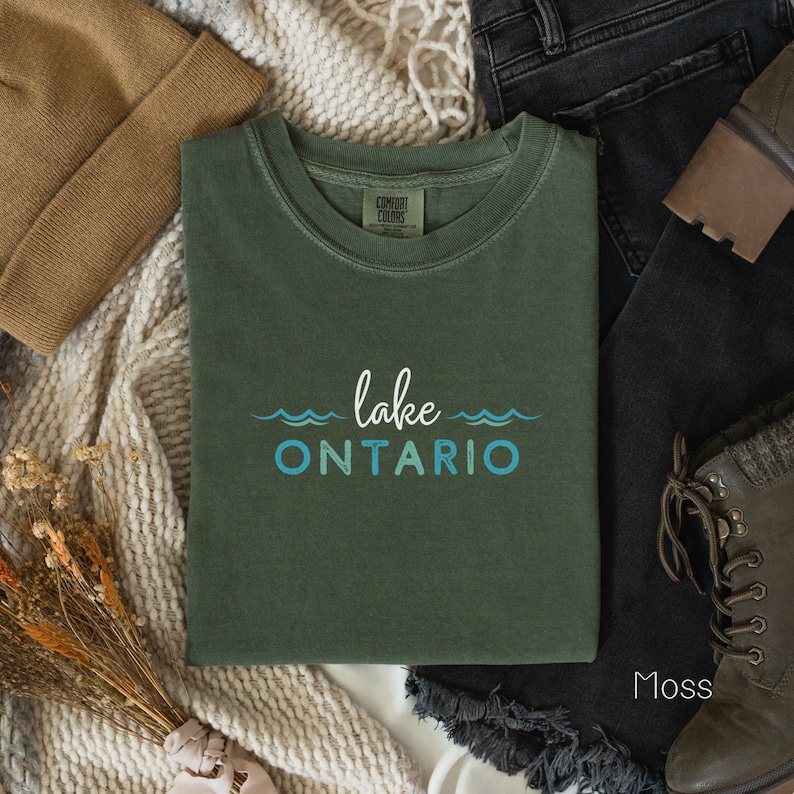 May include: A green t-shirt with the text "lake ONTARIO" in blue and white. The shirt is folded and laid on a white surface with a brown knitted blanket, brown beanie, black jeans, and brown boots.