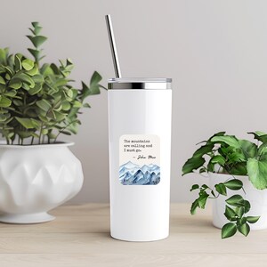 K&ouml;nnte beinhalten: Ein wei&szlig;er Edelstahlbecher mit einem Zitat, das lautet: "Die Berge rufen und ich muss gehen. - John Muir" und einer Aquarellillustration von Bergen.