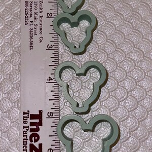May include: Set of five green plastic cookie cutters shaped like Mickey Mouse heads. The cutters are arranged in order of size, with the smallest cutter at the top and the largest cutter at the bottom. The cutters are measured against a ruler, with the largest cutter measuring approximately 6 inches in diameter.