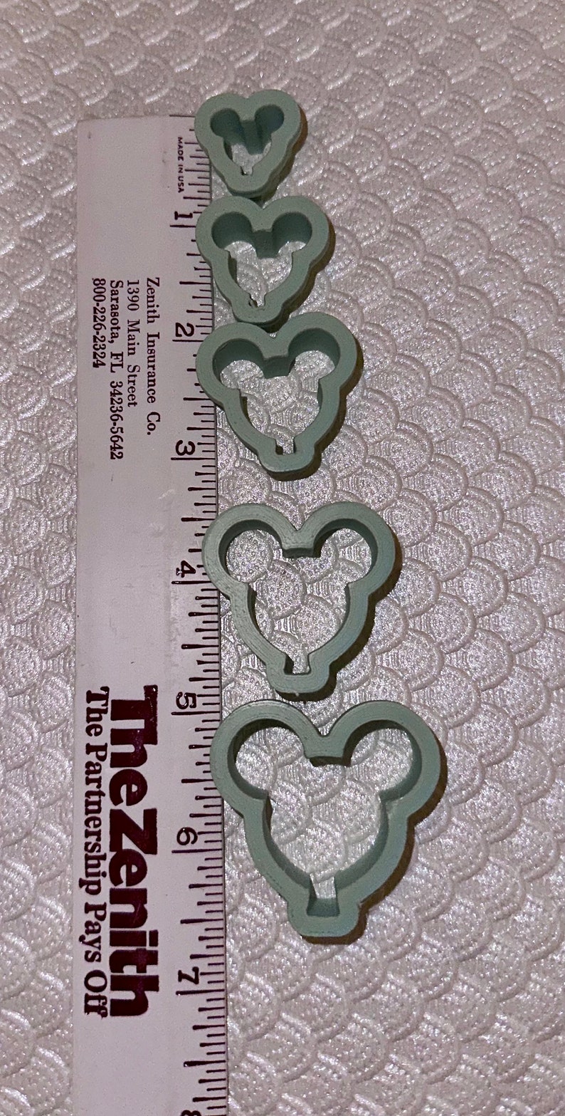 May include: Set of five green plastic cookie cutters shaped like Mickey Mouse heads. The cutters are arranged in order of size, with the smallest cutter at the top and the largest cutter at the bottom. The cutters are measured against a ruler, with the largest cutter measuring approximately 6 inches in diameter.