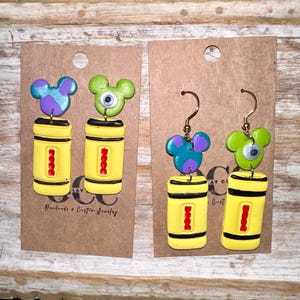 May include: A pair of earrings featuring a yellow crayon with a red stripe and a green monster character with a blue eye. The other earring features a purple monster character with a blue eye. The earrings are handmade and have gold hooks.