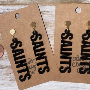 Puede incluir: Un par de pendientes de acrílico negro con postes dorados. Los pendientes tienen la forma de la palabra "Saints".