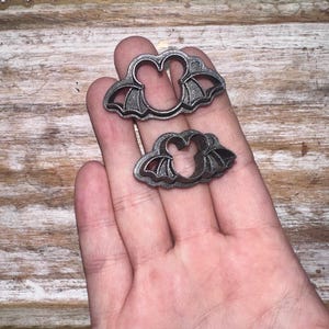 May include: Two metal cookie cutters shaped like bats. The cutters are held in a hand, with the larger cutter on top of the smaller cutter.