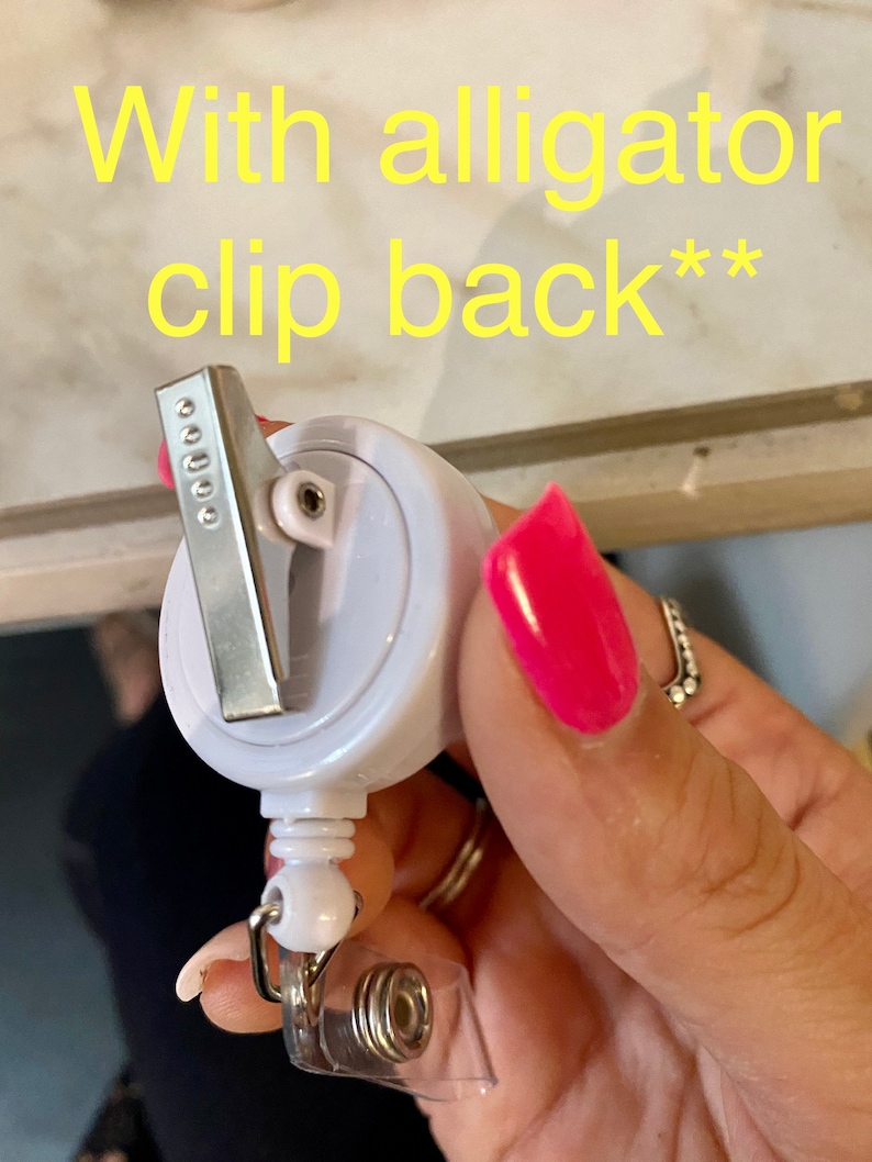 以下が含まれることがあります： 後ろにワニクリップが付いた白いリトラクタブルバッジホルダー。ホルダーは手に持たれています。