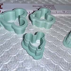 May include: Set of five green plastic cookie cutters shaped like Mickey Mouse heads. The cutters are different sizes.