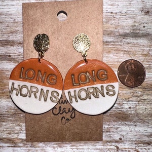 May include: A pair of orange and white earrings with gold accents. The earrings are round and feature the words "Long Horns" in gold lettering. The earrings are made by "Clay Co."