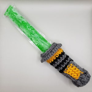 May include: A green popsicle in a clear plastic wrapper is inserted into a crocheted lightsaber holder. The holder is gray, black, and yellow.