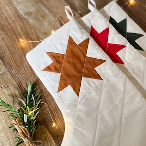 Op de afbeelding: Twee gebroken witte, doorgestikte kerstsokken van stof met grote, veelpuntige sterapplicaties in roestoranje, rood en donkergroen. Elke sok is voorzien van een ophanglus en een doorgestikt ruitpatroon over het gehele oppervlak.
