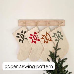 May include: Four cream Christmas stockings with star designs and letters D, C, E, and H. The stockings hang from a wooden rack. The text "paper sewing pattern" is at the bottom of the image. A small evergreen tree is in the lower right corner.