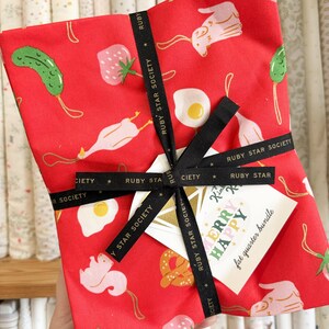 May include: A stack of red fabric featuring a repeating pattern of pink squirrels, green pickles, strawberries, and fried eggs. The fabric is tied with a black ribbon printed with the text Ruby Star Society and includes a tag labelled Merry Happy fat quarter bundle.